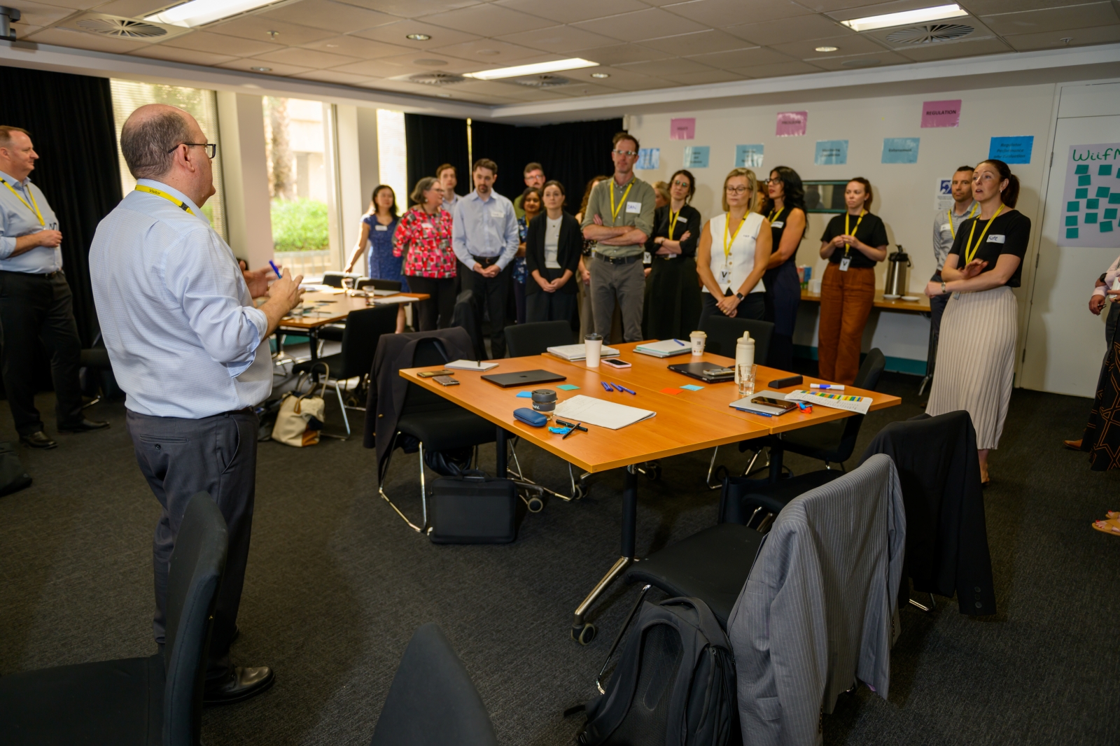 Participants standing in a circle around a table while a facilitator speaks, with issue cards on the wall behind